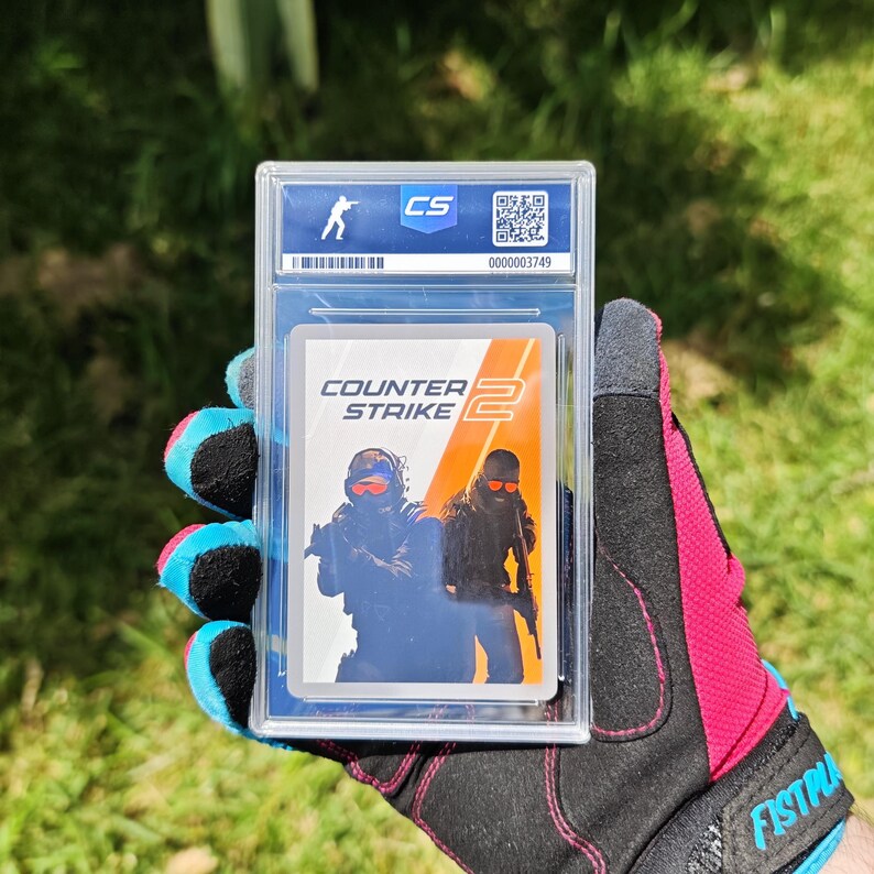 Puede incluir: Una tarjeta coleccionable calificada en una funda protectora transparente. La tarjeta presenta el logotipo "Counter Strike 2" y dos figuras con equipo t&aacute;ctico. La funda tiene un logotipo "CS" y un n&uacute;mero de serie. La tarjeta la sostiene una persona con un guante negro y rosa.