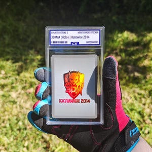 May include: A mint-graded sticker from Counter-Strike 2, featuring 3DMAX (Holo) from Katowice 2014. The sticker is encased in a clear protective holder. The sticker has a red and yellow graphic design with the text "KATOWICE 2014".