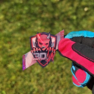 K&ouml;nnte beinhalten: Ein Aufkleber mit einem roten Teufelslogo, Kopfh&ouml;rern und den Buchstaben "BD" in einem Schilddesign. Der Aufkleber wird von einem rot-schwarzen Handschuh mit blauen Akzenten vor einem gr&uuml;nen, grasbewachsenen Hintergrund gehalten.