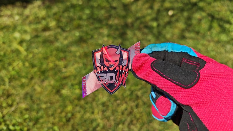 K&ouml;nnte beinhalten: Ein Aufkleber mit einem roten Teufelslogo, Kopfh&ouml;rern und den Buchstaben "BD" in einem Schilddesign. Der Aufkleber wird von einem rot-schwarzen Handschuh mit blauen Akzenten vor einem gr&uuml;nen, grasbewachsenen Hintergrund gehalten.