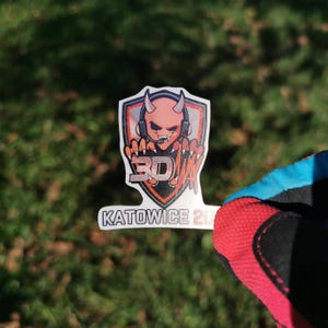 May include: A sticker featuring a devil character with headphones, horns, and a microphone. The design includes the text "3D" and "Katowice 20". The sticker is white with orange, red, and black accents, set against a green background.