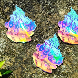 Könnte beinhalten: Drei regenbogenfarbene holografische Aufkleber mit einem abstrakten Design, das einer Flamme ähnelt. Die Aufkleber befinden sich auf einer rauen, strukturierten Oberfläche.