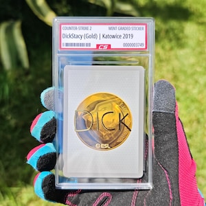 Puede incluir: Una pegatina de Counter-Strike 2 de grado superior, con "DickStacy (Gold) | Katowice 2019". La pegatina est&aacute; dentro de una funda protectora transparente. La pegatina tiene un dise&ntilde;o circular dorado con la palabra "DICK".