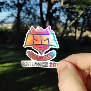 Peut inclure: Un autocollant holographique avec le logo Hellraisers, présentant un design rouge, orange et jaune avec le texte "Hellraisers" et "Katowice 2019".