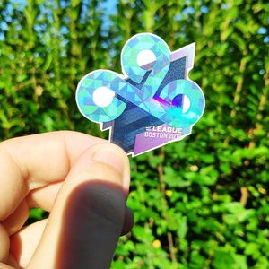 May include: A sticker featuring a geometric design in shades of blue, green, and purple, with the text "E LEAGUE BOSTON 2018". The sticker is held against a backdrop of green foliage.