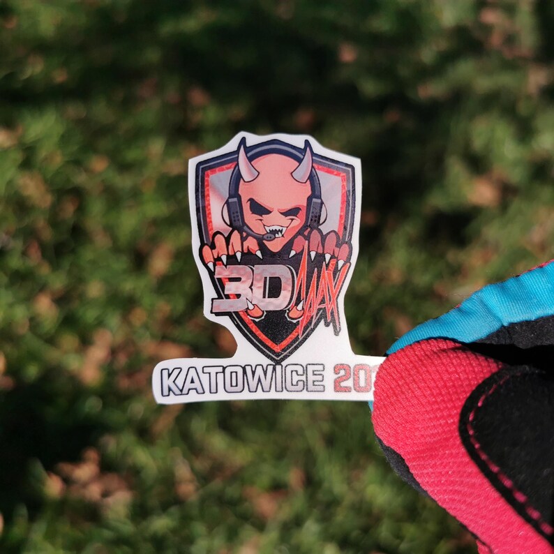 May include: A sticker featuring a cartoon devil with headphones, horns, and sharp claws. The devil is inside a shield with the text "3D JAY" and "KATOWICE 20". The sticker is held by a person wearing a red and blue glove.