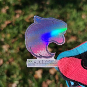 May include: A holographic sticker in the shape of a stylized animal head, with iridescent colors of blue, green, and pink. The sticker has the text "COPENHAGEN 20" at the bottom. The sticker is held by a person wearing a red and black glove.
