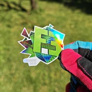 Può includere: Un adesivo colorato con un design geometrico e la lettera "F" in verde. L'adesivo ha un effetto olografico e il testo "COLOGNE 2016". L'adesivo è tenuto da una persona che indossa un guanto rosa e nero.