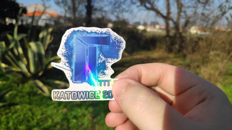 May include: A holographic sticker with a blue and white design. The sticker features a stylized letter "T" and the text "KATOWICE 2020".