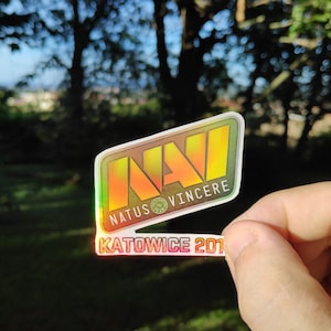 Puede incluir: Una pegatina holográfica con el texto "NAVI NATUS VINCERE KATOWICE 2019" en un esquema de colores amarillo, naranja y verde.