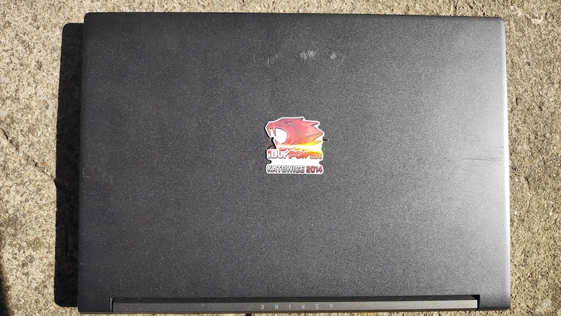 May include: A black laptop with a red, orange, yellow, and green sticker on the lid. The sticker features a cartoon red dragon with the text "LEAGUE POWER" and "KNOWWIDE 2014".