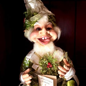 Puede incluir: Una caprichosa figura de elfo navideño con una cara sonriente, barba blanca y un sombrero alto y adornado. El elfo viste de verde y blanco con detalles decorativos, sosteniendo un certificado. Para decoración navideña.