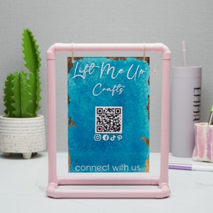 May include: Pink PVC pipe frame holding a blue and gold watercolor sign with the text "Lift Me Up Crafts" and a QR code. The sign also includes social media icons and the text "connect with us".