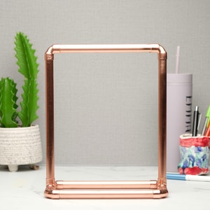 May include: A copper pipe shelf with a rectangular frame. The shelf is empty and is sitting on a white surface.