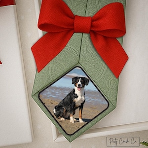 May include: A decorative ornament featuring a photo of a dog on a beach, set within a green diamond-shaped frame. A large red bow is attached to the top. The ornament has a textured pattern.