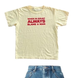 May include: A pale yellow t-shirt with the red text "WHEN IN DOUBT ALWAYS BLAME A MAN", combined with blue denim shorts. The shorts feature a button fastening and pockets.