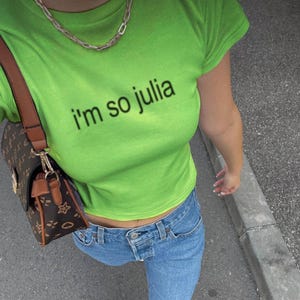 May include: A lime green cropped t-shirt with the text "i'm so julia" printed on the front. The shirt is being worn with blue jeans and a brown and black handbag.