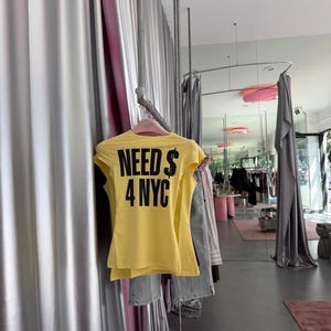 May include: A bright yellow short-sleeved t-shirt hanging on a pink hanger. The back of the shirt features bold black text that reads "NEED $ 4 NYC" in a sans-serif font. The garment is displayed on a metal clothing rack in a retail space.