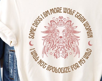 May include: A cream-colored t-shirt with a brown and pink graphic of a wolf with floral details. The text "Some days I am more wolf than woman I will not apologize for my wild" is printed in a circle around the wolf graphic.