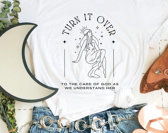 May include: White t-shirt with black text and a line drawing of a woman with a halo. The text reads "Turn it over to the care of God as we understand her."