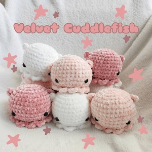 May include: Six crocheted plush cuddlefish toys in pink and white velvet. The toys are arranged in a circle on a white fluffy surface with pink stars scattered around them. The text "Velvet Cuddlefish" is written in pink letters above the toys.