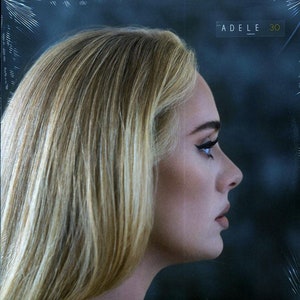 May include: A close-up of a woman's face in profile, with blonde hair and dark eyeliner. The background is a dark blue. The text "ADELE 30" is visible in the upper right corner.