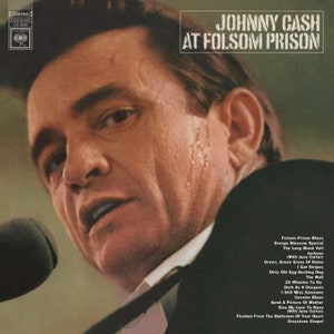 May include: A black and white photo of Johnny Cash performing live at Folsom Prison. The image shows Cash singing into a microphone with a serious expression on his face. The album title "Johnny Cash At Folsom Prison" is printed in white letters on a green background at the top of the image. The album's tracklist is printed in white text on a black background at the bottom of the image.
