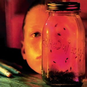 May include: A glass jar with a metal lid, filled with a red liquid and small black insects. The words "Alice in Chains" are etched on the jar. A person's face is visible in the background, illuminated by red light.