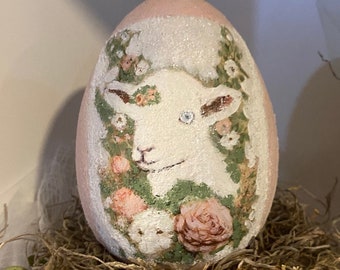 Puede incluir: Un huevo de Pascua rosa con un cordero blanco pintado en él, rodeado de flores rosas y blancas. El huevo está sentado en un nido de paja marrón.