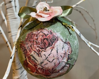 May include: A green and pink Christmas ornament with a floral design. The ornament is decorated with a pink flower and a green ribbon.