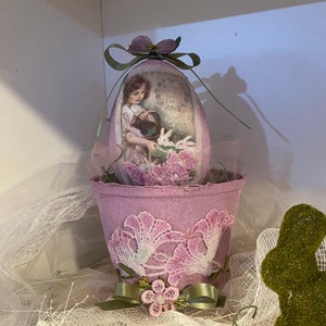 Puede incluir: Un huevo de Pascua de tela morada con una imagen vintage de una niña y conejos, anidado en una maceta de tela morada con ribete de encaje y una cinta verde. La maceta está decorada con flores rosas.