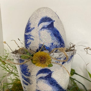 Puede incluir: Un huevo de cerámica blanca con un diseño de pájaro azul y detalles de purpurina se coloca en una pequeña maceta de cerámica blanca con un diseño de pájaro azul y detalles de purpurina. La maceta está decorada con una flor amarilla y una cuerda.