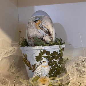 Puede incluir: Una maceta de cerámica blanca con un pájaro azul y hojas verdes pintadas en el lateral. Un huevo azul con un pájaro pintado en él se encuentra dentro de la maceta.