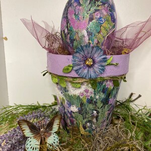 May include: A purple flower pot with a floral design and a purple egg with a floral design on top. The pot is decorated with a purple flower and green leaves. The pot is sitting on a bed of moss.