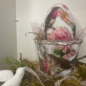 Puede incluir: Un huevo de Pascua decorativo con un diseño de pájaro y flores, colocado en una maceta a juego. El huevo y la maceta están adornados con purpurina y se asientan sobre un lecho de musgo, con una figura de pájaro blanco y una cinta cerca.