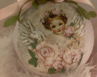 May include: A round, light pink ornament decorated with a vintage-style image of a cherub holding roses. The ornament is adorned with a pink ribbon, a pink fabric rose, and green leaves. The ornament is resting on a bed of white feathers.