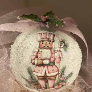 Peut inclure: Une boule de Noël ronde et blanche avec une surface texturée. L'ornement présente une image peinte d'un soldat casse-noisette rose et blanc. Un ruban rose et du houx vert avec des baies roses ornent le dessus.