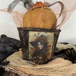 May include: A small, rustic, black metal planter with a burlap-wrapped pumpkin inside. The planter has a painted image of a witch with a black hat and long dark hair. The planter is decorated with a black ribbon and a sheer peach-colored fabric.