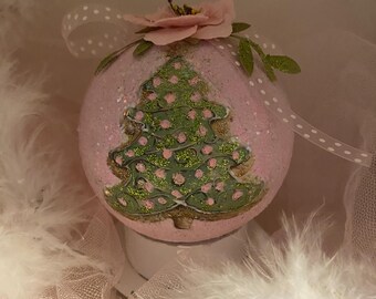 May include: A pink Christmas ornament with a glittery finish. The ornament features a green Christmas tree design with pink polka dots. A pink flower and green leaves adorn the top, with a white polka dot ribbon for hanging. The ornament is surrounded by white feathers.