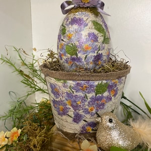 Puede incluir: Un huevo de Pascua decorativo con un diseño floral morado y purpurina está anidado en una maceta pequeña con un diseño floral similar. Un pájaro plateado brillante se sienta junto a la maceta.
