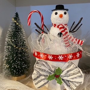 Könnte beinhalten: Festliche Schneemann-Dekoration mit schwarzem Zylinder, Zweigarmen und einem rot-weiß gestreiften Schal. Der Schneemann befindet sich in einem weißen Topf mit rotem Schneeflockenband und einer Noten-Schleife. Ein kleiner Weihnachtsbaum und eine Zuckerstange ergänzen die Szene.