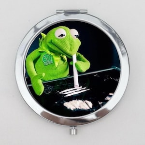 May include: A round, silver-framed compact mirror with a black and white image of a green frog puppet holding a white straw and a white powder on a black surface. The frog is wearing a green shirt with a green "30" on it.