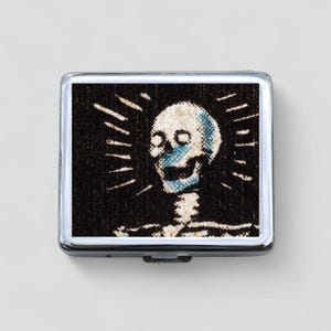 Peut inclure: Un étui à cigarettes en argent avec une image en noir et blanc d'un crâne avec un visage bleu. Le crâne est entouré d'une auréole de lumière blanche.