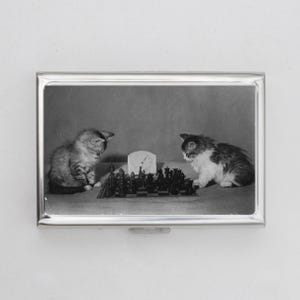 Pode incluir: Uma fotografia a preto e branco de dois gatinhos a jogar xadrez. Os gatinhos estão sentados numa mesa com um tabuleiro de xadrez à sua frente. Um gatinho está a olhar para o tabuleiro de xadrez, enquanto o outro está a olhar para a câmara.