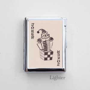 May include: A rectangular silver-toned metal lighter featuring a cream-colored inlay on the front. The inlay displays a black line drawing of a clown popping out of a jack-in-the-box with the word JOKER printed vertically on both sides.