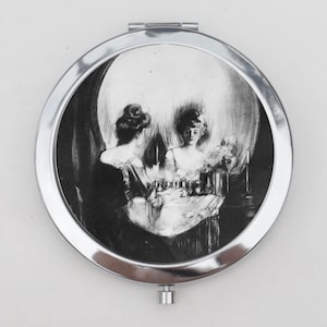 May include: A round, silver-toned compact mirror featuring a black and white illustration of a woman sitting at a vanity table. The arrangement of the woman, her reflection, and the surrounding shadows creates an optical illusion of a human skull.