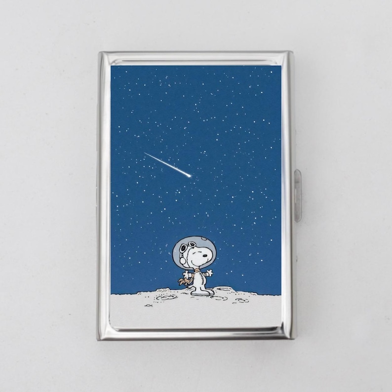 K&ouml;nnte beinhalten: Ein silbernes Zigarettenetui mit blauem Hintergrund, auf dem ein Cartoon-Hund in einem Astronautenanzug und ein Sternschnuppen zu sehen sind. Der Hund steht auf einer wei&szlig;en Oberfl&auml;che mit Kratern.