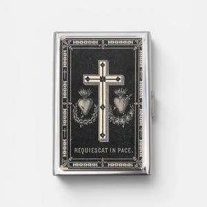 May include: A rectangular silver-toned metal case featuring a black inlay with a central white cross, two sacred hearts, and the text "REQUIESCAT IN PACE." The design is framed by a decorative border with repeating "X" patterns and small crosses.