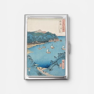 May include: A rectangular metal cigarette case featuring a reproduction of a Japanese woodblock print. The artwork depicts a coastal bay with blue water, several sailing boats, green hills, and mountains, including Japanese text characters.