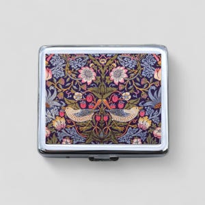 May include: A silver metal pill box with a floral design featuring birds and berries. The design is in shades of blue, green, pink, and white on a dark purple background.